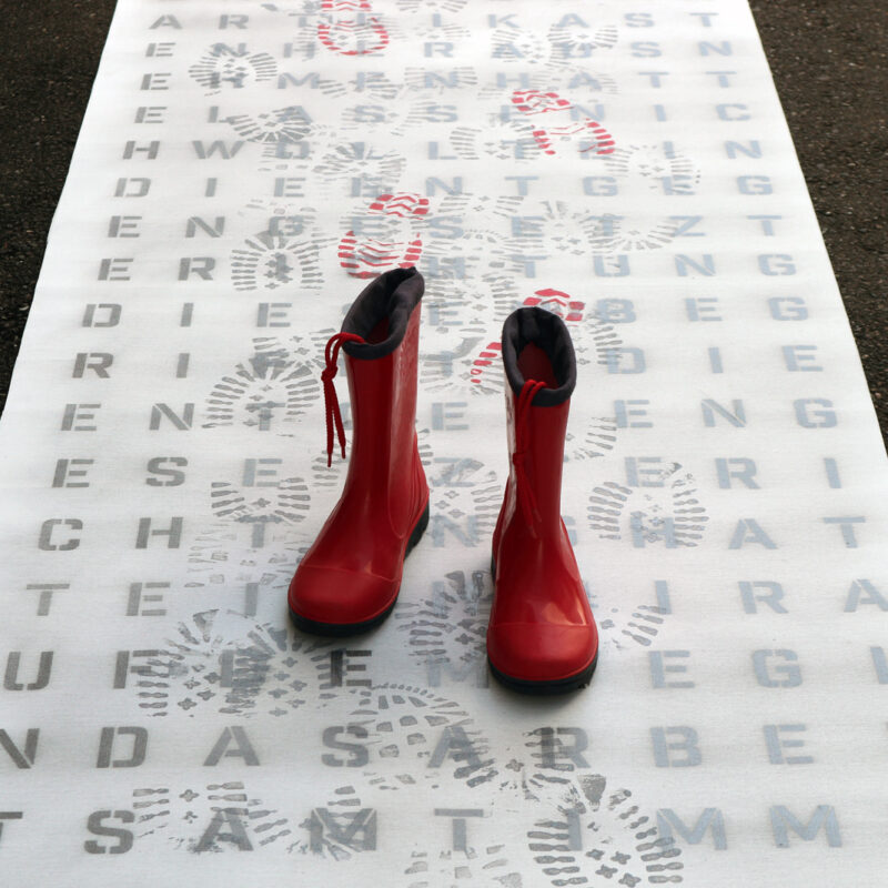 Ein Paar rote Gummistiefel auf einer Leinwand mit Buchstaben und den Abdrücken von Schuhen